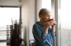 A mature woman with dementia sips her coffee alone while staring out her window.