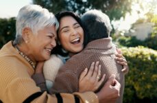 A young adult smiles as they are sandwiched in a hug between their older parents while outdoors on a sunny day