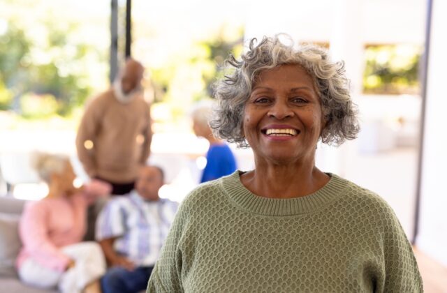 A smiling older woman in an assisted living home. A group of other seniors are blurred in the background.