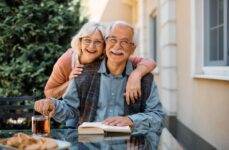 A senior woman hugging her husband from behind and smiling while sitting at an outdoor patio table in assisted living.