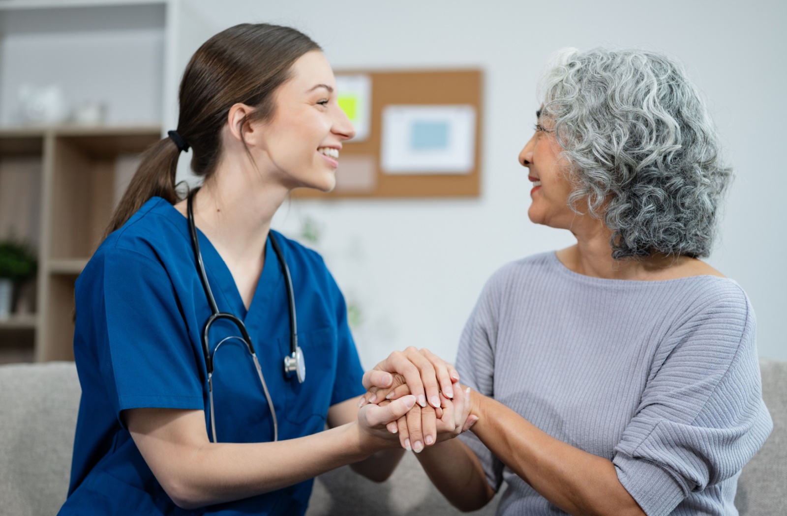 A caregiver and a senior woman clasping hands and smiling at each other while sitting on a couch in assisted living.
