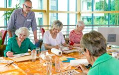 A group of focused seniors taking an art class use watercolors to paint pictures in a warm, sunny room.