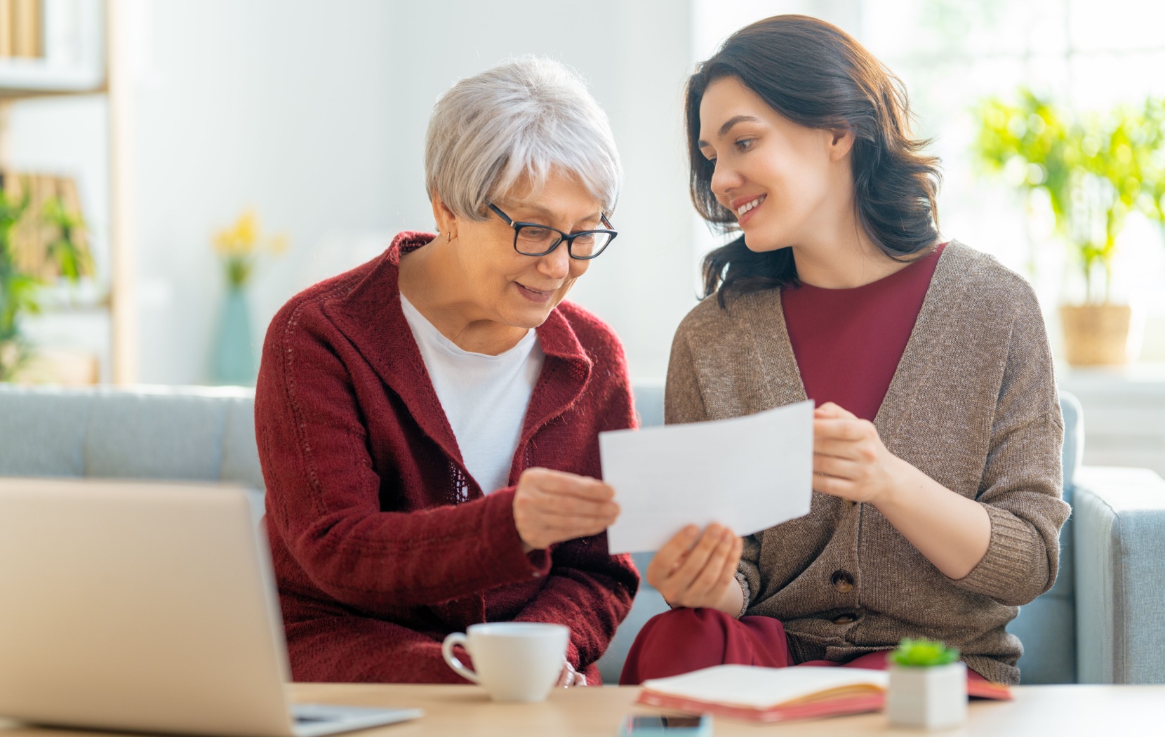 A senior and their adult child smile and reminisce while they look at old pictures and journals together and drink tea.