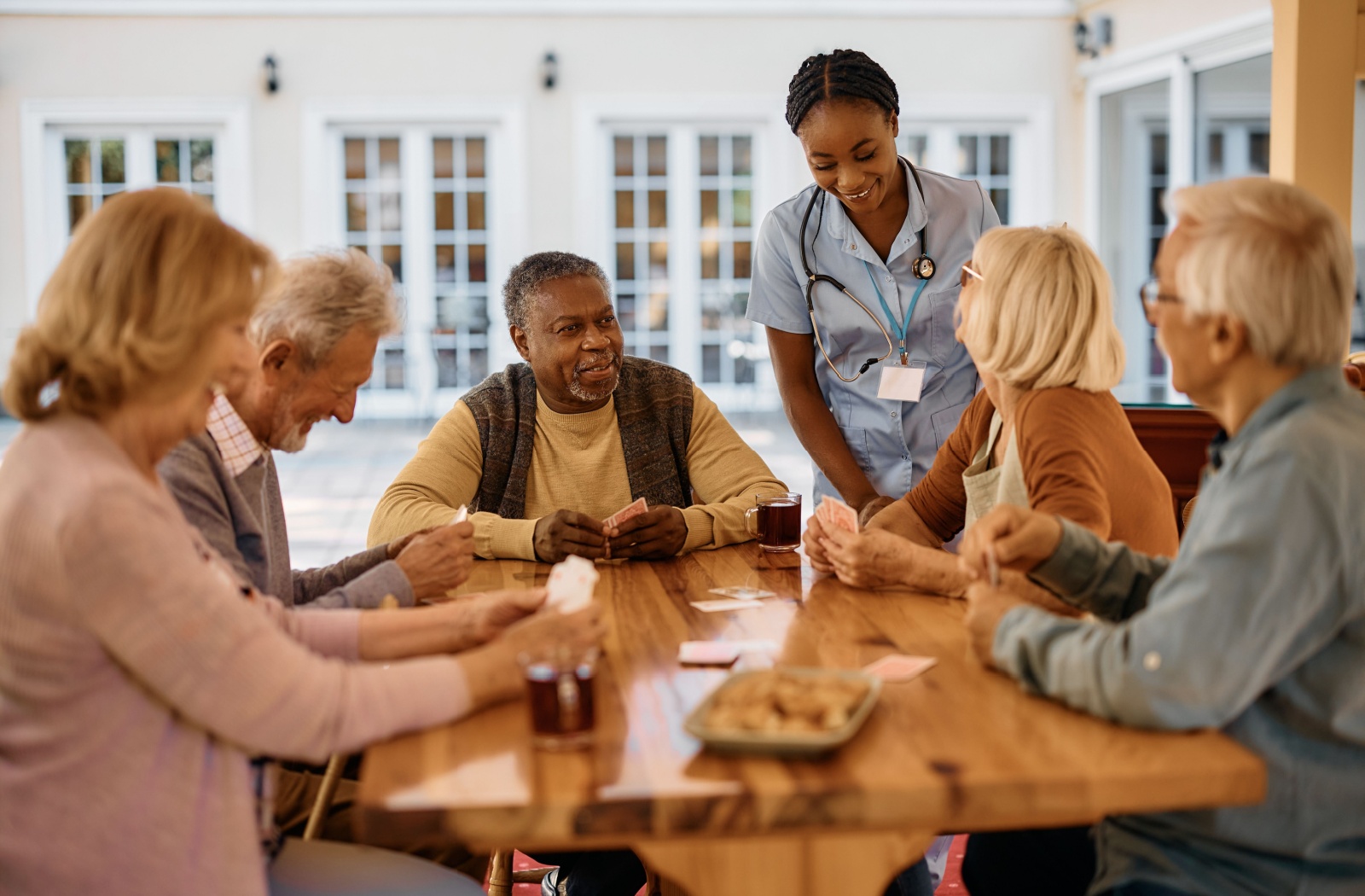 A group of seniors sit around an outdoor table playing cards and having drinks while a nurse checks in on them.